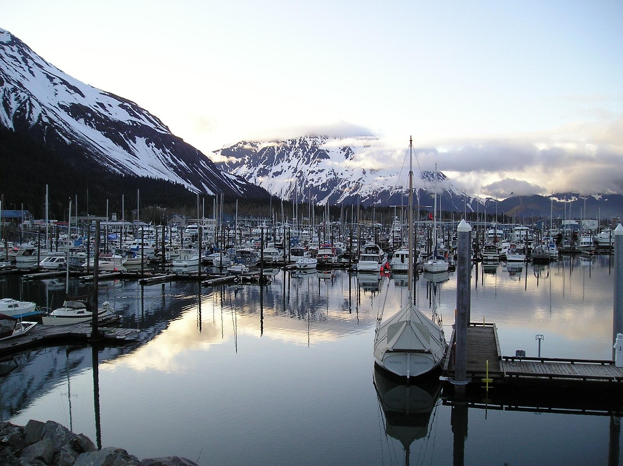 Alaska harbor marina filled with boats and yachts with snow-covered mountains reflected in calm water.