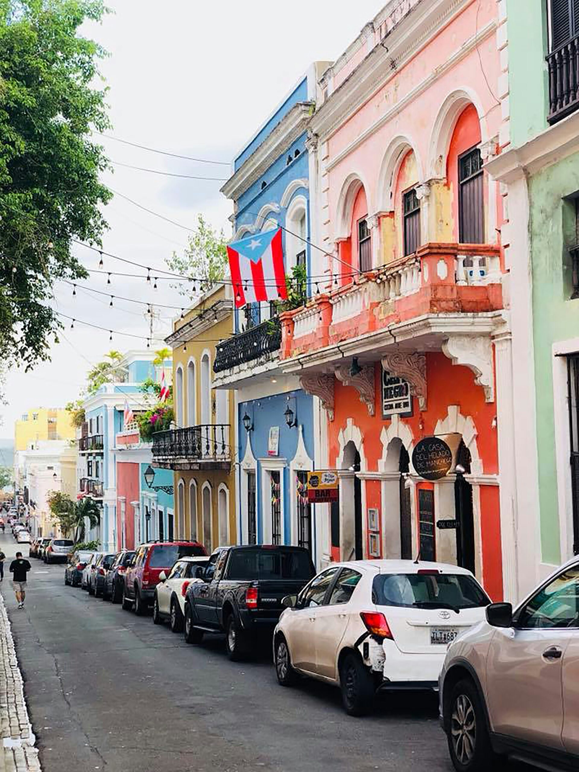 Colorful historic street in Old San Juan, Puerto Rico, with parked cars and a Puerto Rican flag hanging from a balcony.