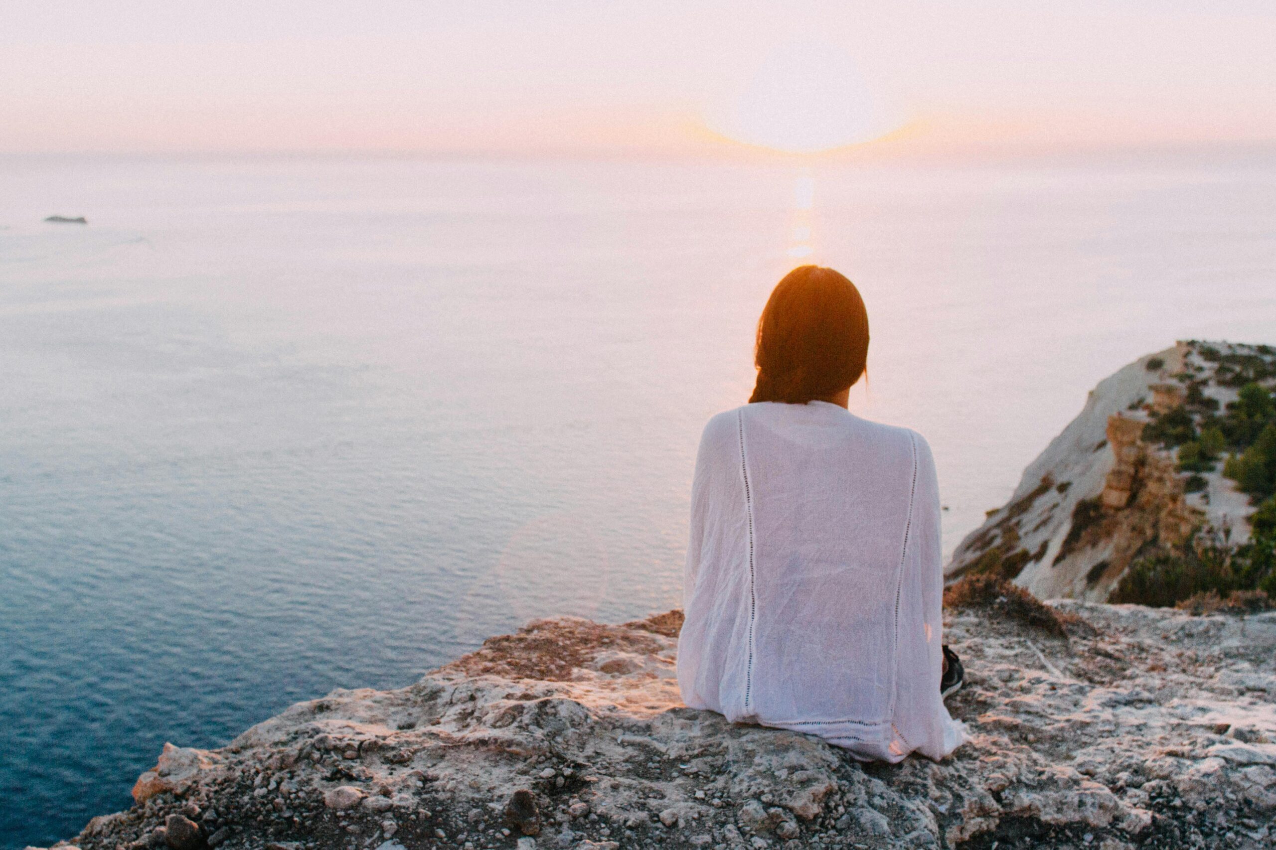 Woman sitting on a rocky cliff looking out at a calm ocean sunset, symbolizing a peaceful fresh start after moving.
