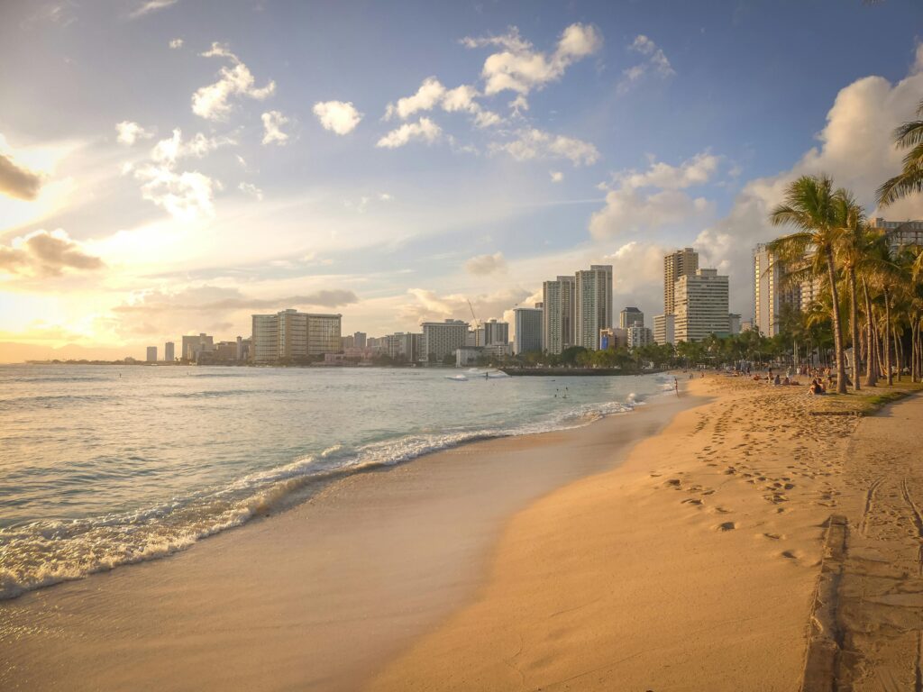Golden hour view of a sandy beach in Hawaii with palm trees, gentle waves, and a modern city skyline in the background.