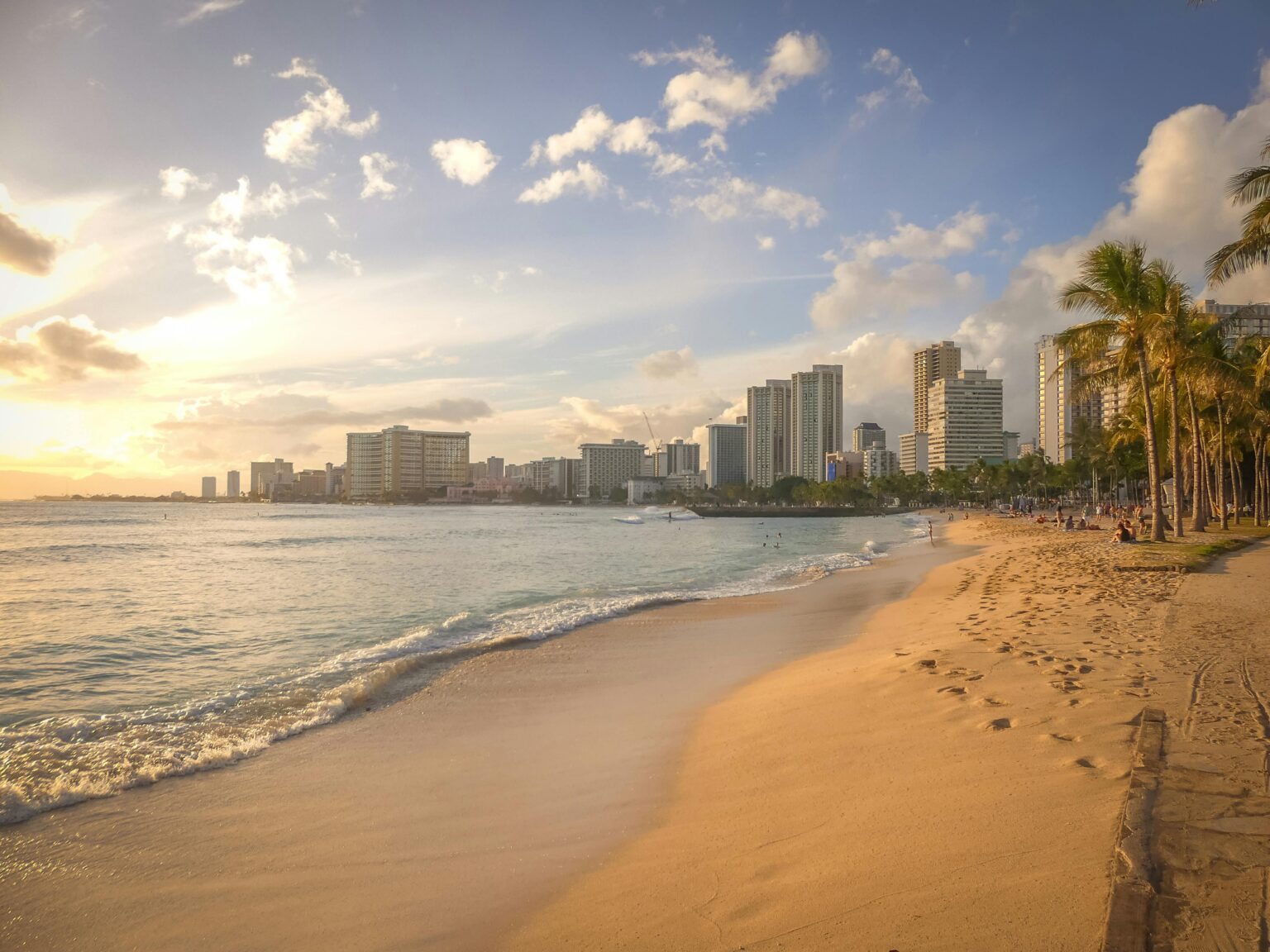 Golden hour view of a sandy beach in Hawaii with palm trees, gentle waves, and a modern city skyline in the background.