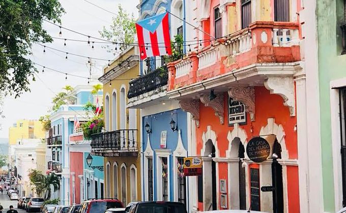Colorful historic street in Old San Juan, Puerto Rico, with parked cars and a Puerto Rican flag hanging from a balcony.
